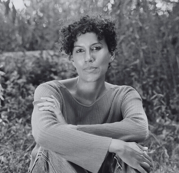 A black-and-white portrait of Aracelis Girmay sitting on the grass with tall plants behind her. Een zwart-witportret van Aracelis Girmay zittend op het gras met hoge planten achter haar.