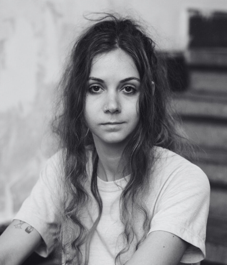A black-and-white portrait of Dominique De Groen on the stairs, looking kindly into the camera. Een zwart-witportret van Dominique de Groen op de trap, die vriendelijk in de camera kijkt.