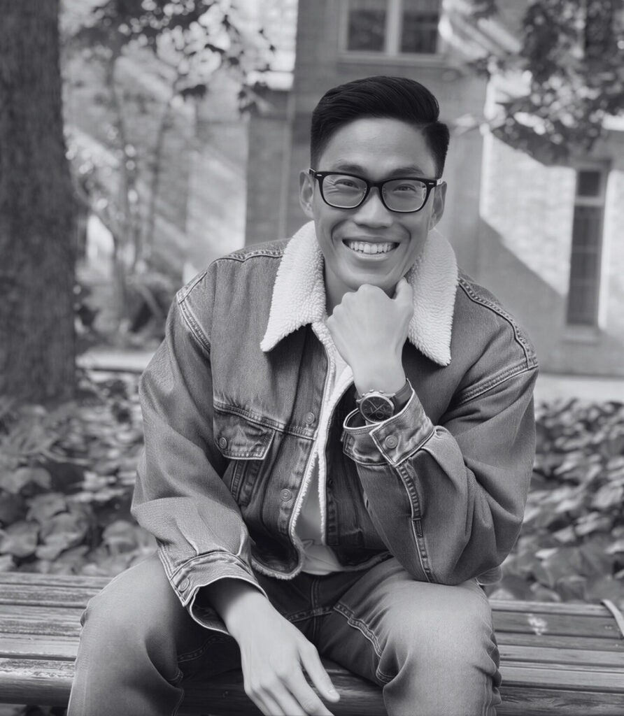 A black-and-white picture of Hendri Yulius Wijaya on a bench with a tree and building in the background. Een zwart-witfoto van Hendri Yulius Wijaya op een bank met een boom en een gebouw op de achtergrond.