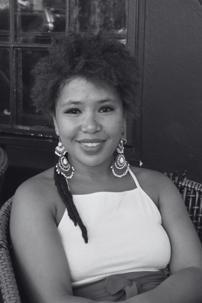 A black-and-white picture of Lynthia Julius on a wicker chair by a window in a dark wall. Een zwart-witfoto van Lynthia Julius op een rieten stoel bij een raam in een donkere muur.