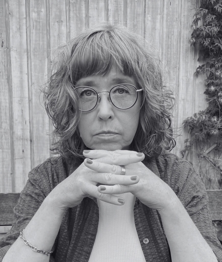 A black-and-white photo of Aase Berg looking directly at the camera, her hands clasped together. Een zwart-witfoto van Aase Berg die recht in de camera kijkt, met haar handen in elkaar gevouwen.