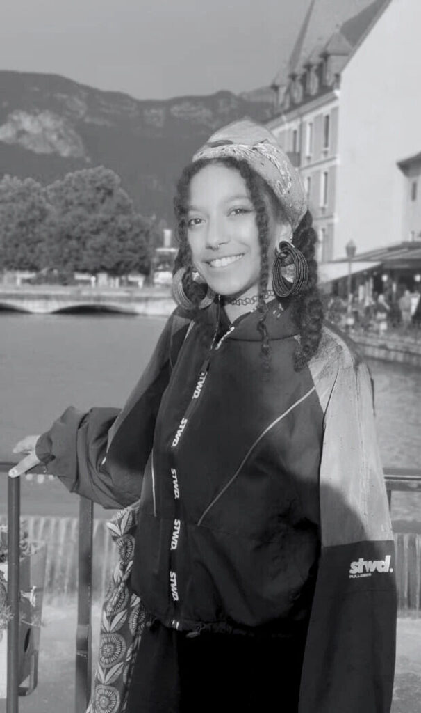 A black-and-white photo of Bakita Pietrowicz standing on a bridge overlooking a river, with mountains and a building in the background. Een zwart-witfoto van Bakita Pietrowicz op een brug boven een rivier, met bergen en een gebouw op de achtergrond.