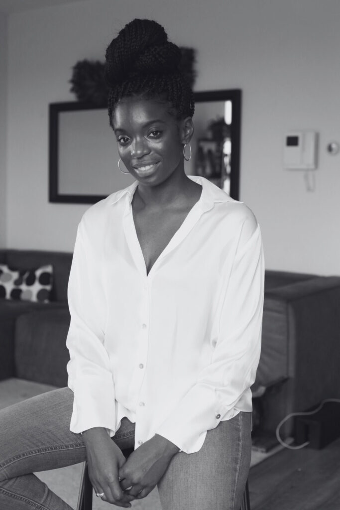A black-and-white picture of Yahmani Blackman sitting on a stool in a living room. Een zwart-witfoto van Yahmani Blackman die op een kruk zit in een woonkamer.
