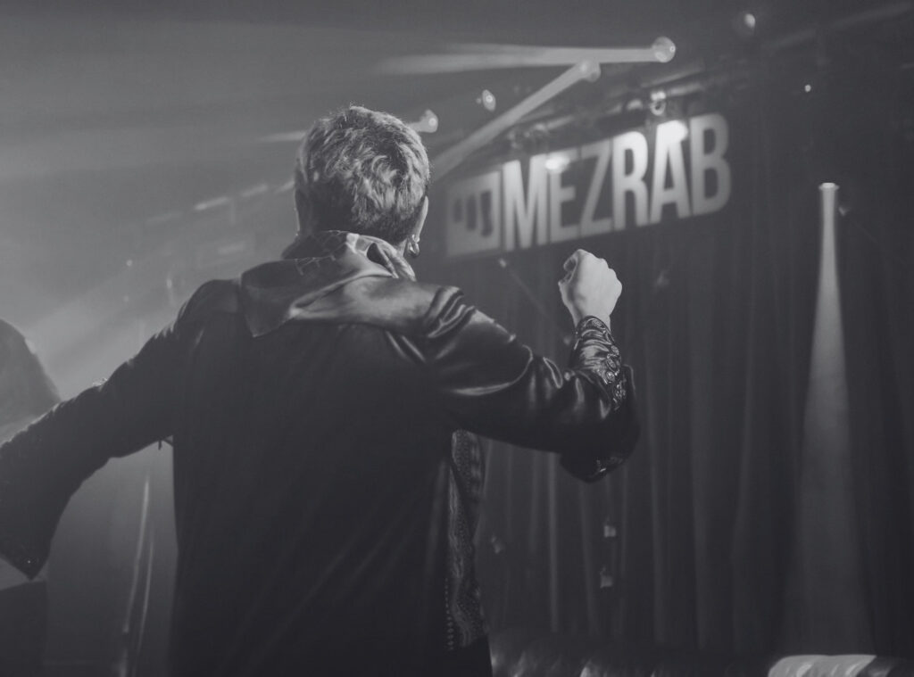 A black-and-white picture of a person dancing with 'MEZRAB' in the background. Een zwart-witfoto van een persoon die aan het dansen is met op de achtergrond 'MEZRAB'.