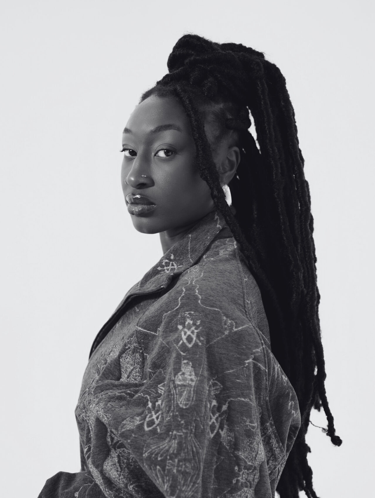 A black-and-white portrait of Ella John shot from the side, with their head turned toward the camera. Een zwart-witportret van Ella John, van opzij gefotografeerd, met het hoofd naar de camera gedraaid.