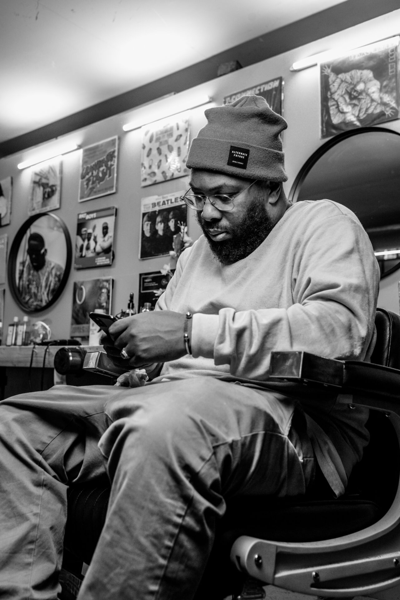 A black-and-white photo of Ivan Winter sitting in a barber’s chair, looking at his phone. Een zwart-witfoto van Ivan Winter die in een kappersstoel zit en op zijn telefoon kijkt.