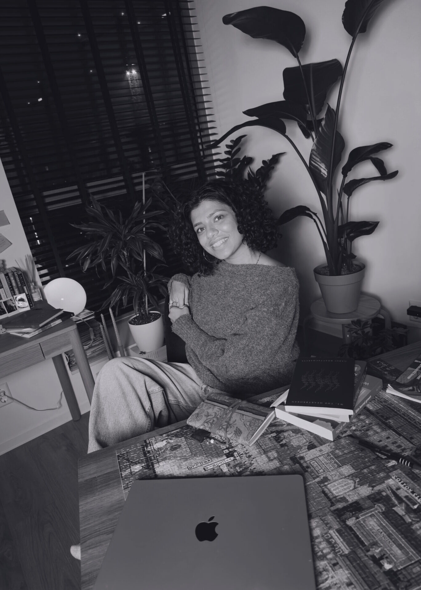 A black-and-white picture of Charisa Chotoe smiling and sitting at a table with books and a laptop on it. Een zwart-witfoto van Charisa Chotoe die glimlacht en aan een tafel zit met boeken en een laptop erop.