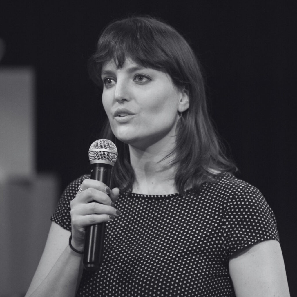 A black-and-white picture of Kila van der Starre holding a microphone on stage. Een zwart-witfoto van Kila van der Starre die een microfoon vasthoudt op het podium.