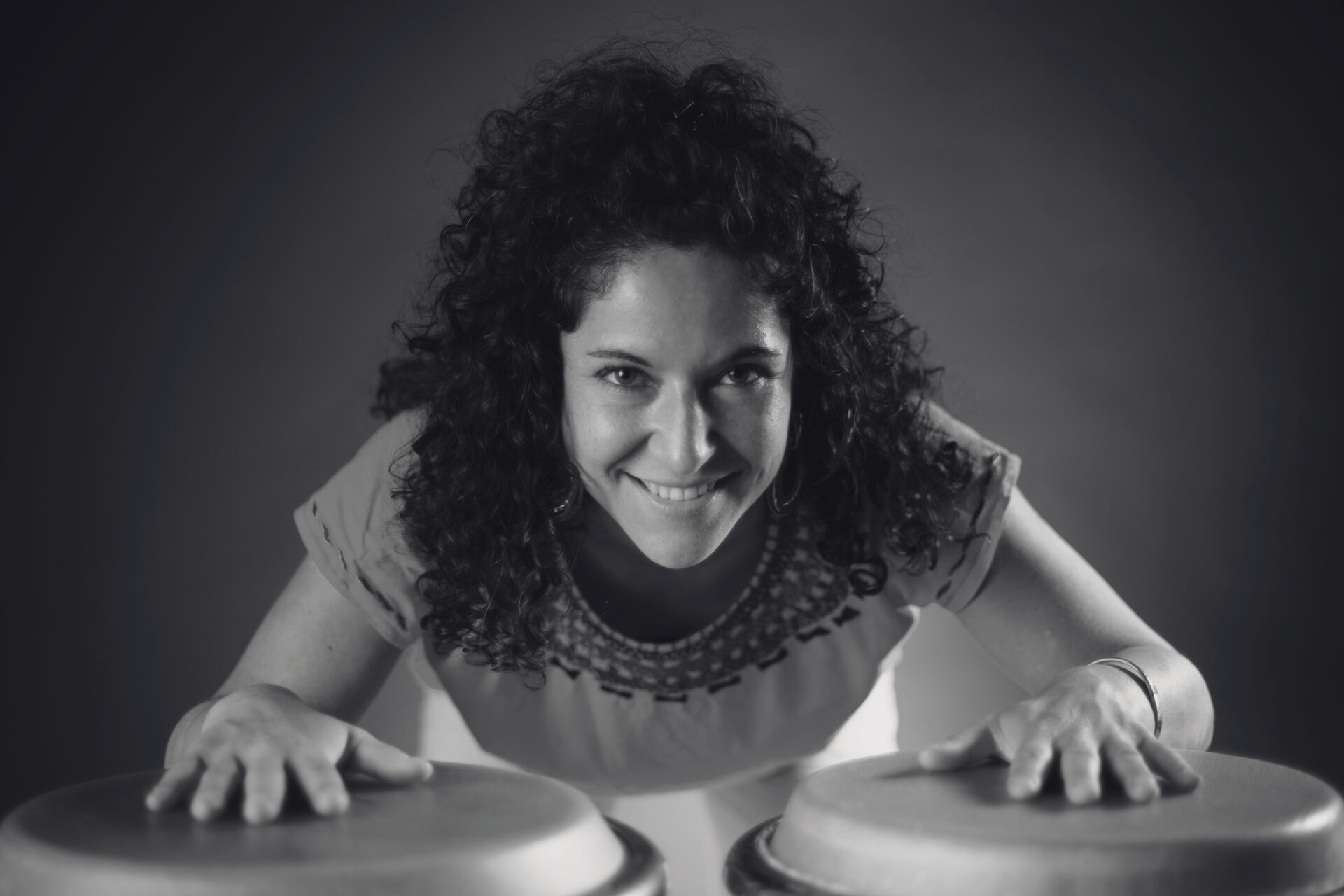 A black-and-white picture of Lauriane Ghils with her hands on percussion instruments, smiling at the camera. Een zwart-witfoto van Lauriane Ghils die lacht naar de camera met haar handen op percussie-instrumenten.