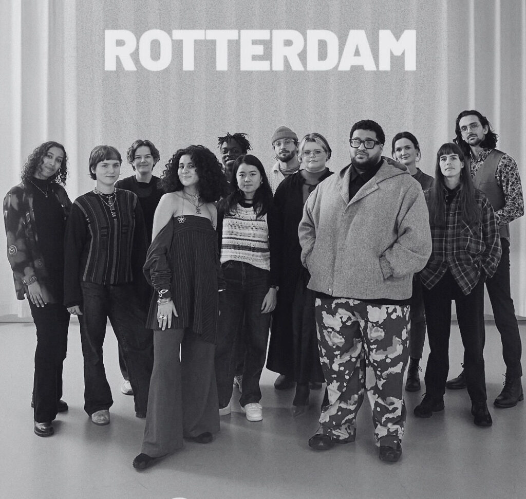 A black-and-white photo of a group of people posing in front of a sign that says ‘Rotterdam. Een zwart-witfoto van een groep mensen die poseert voor een ‘Rotterdam’-sign.